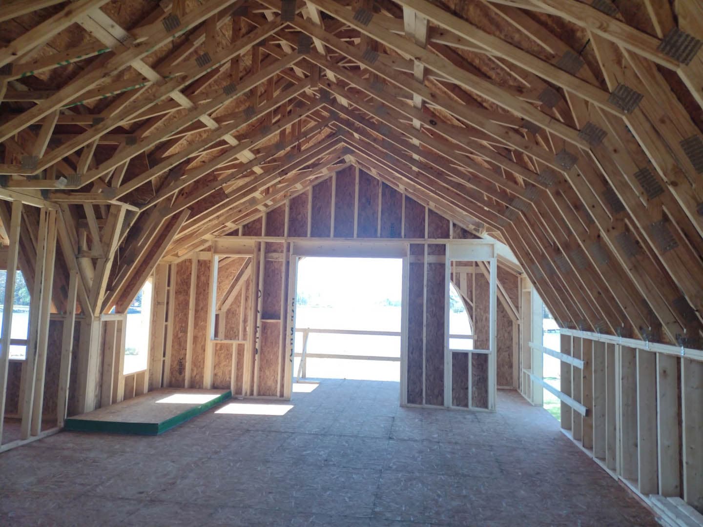 Interior construction photograph of a barn showing an exposed gambrel-style roof truss system with diagonal knee braces, OSB sheathing, and a large open doorway framing a pastoral view