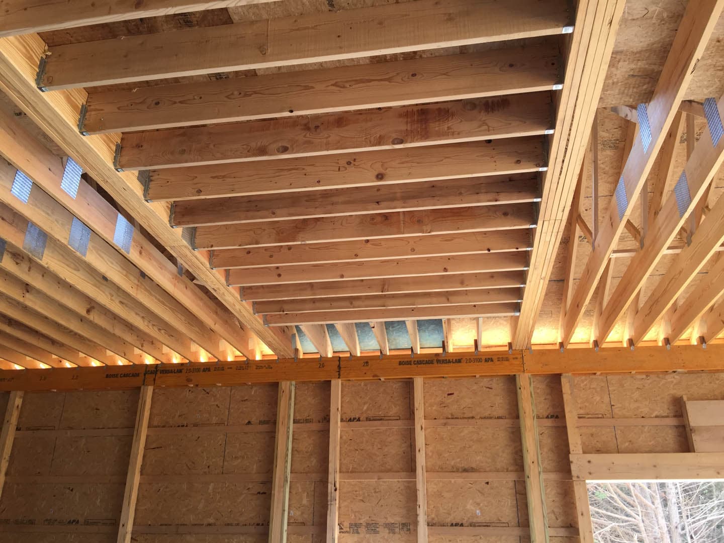 Construction photograph looking up at exposed roof framing inside a barn building, showing parallel dimensional lumber rafters and engineered I-joists with OSB sheathing
