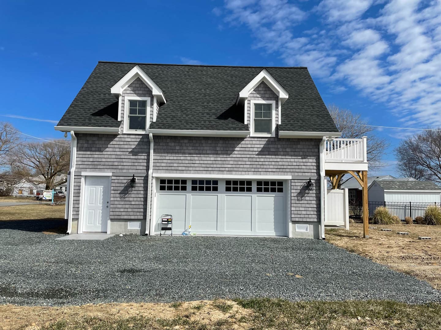 Newly constructed two-car garage with living space above featuring gray cedar shake siding, two dormer windows, a dark shingle roof, and a small second-floor deck with timber pergola