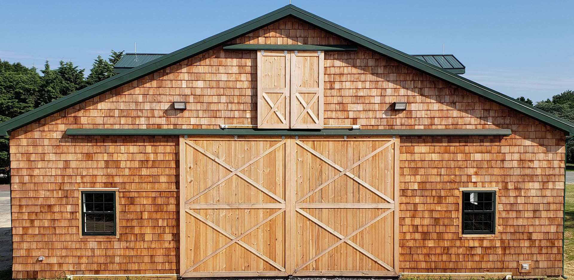 Exterior of a cedar shingle barn facade with a large pair of sliding X-brace wooden doors, a second-level hayloft door, flanking windows, and green metal roof trim