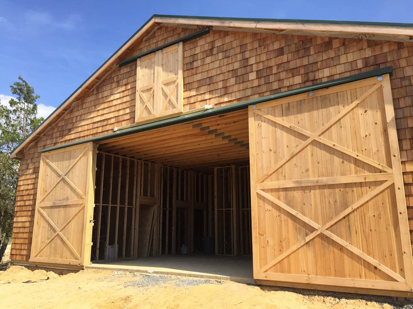 Exterior construction photograph of a cedar shingle barn with large sliding X-brace wooden doors open, revealing exposed interior wood stud framing