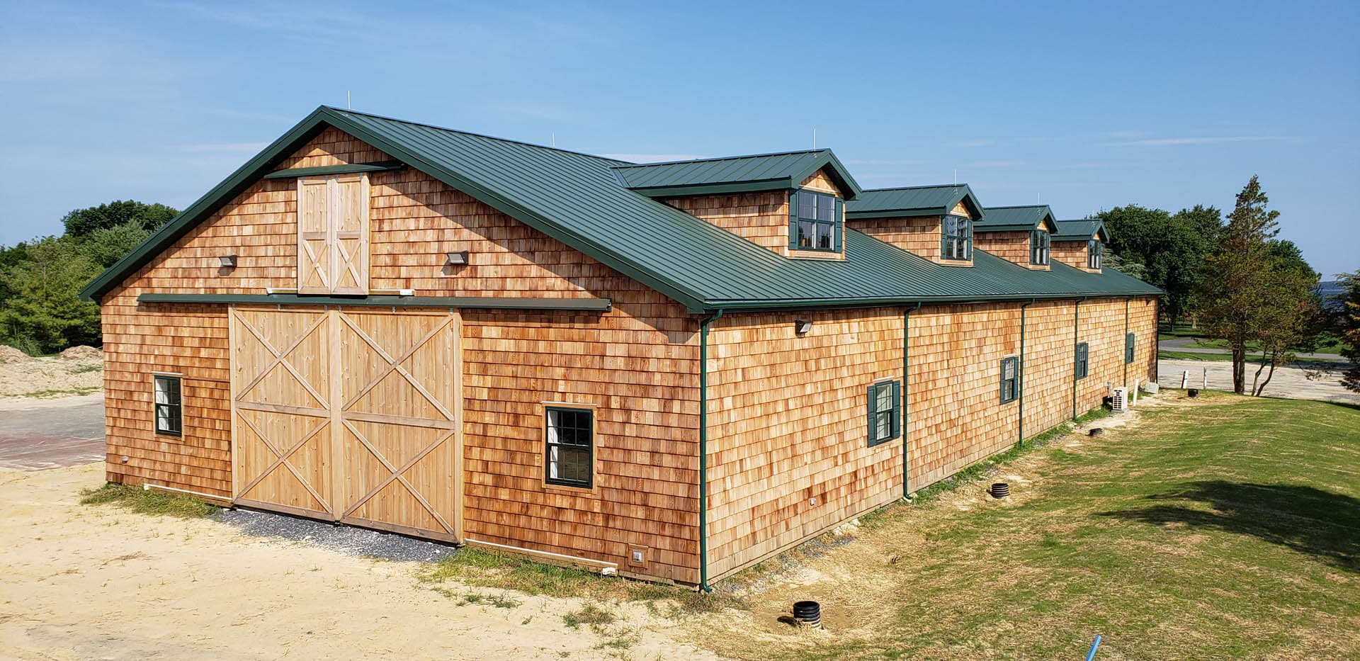 Newly constructed cedar shingle equestrian barn with a green standing-seam metal roof, large sliding barn doors with X-brace detail, dormer windows, and green trim