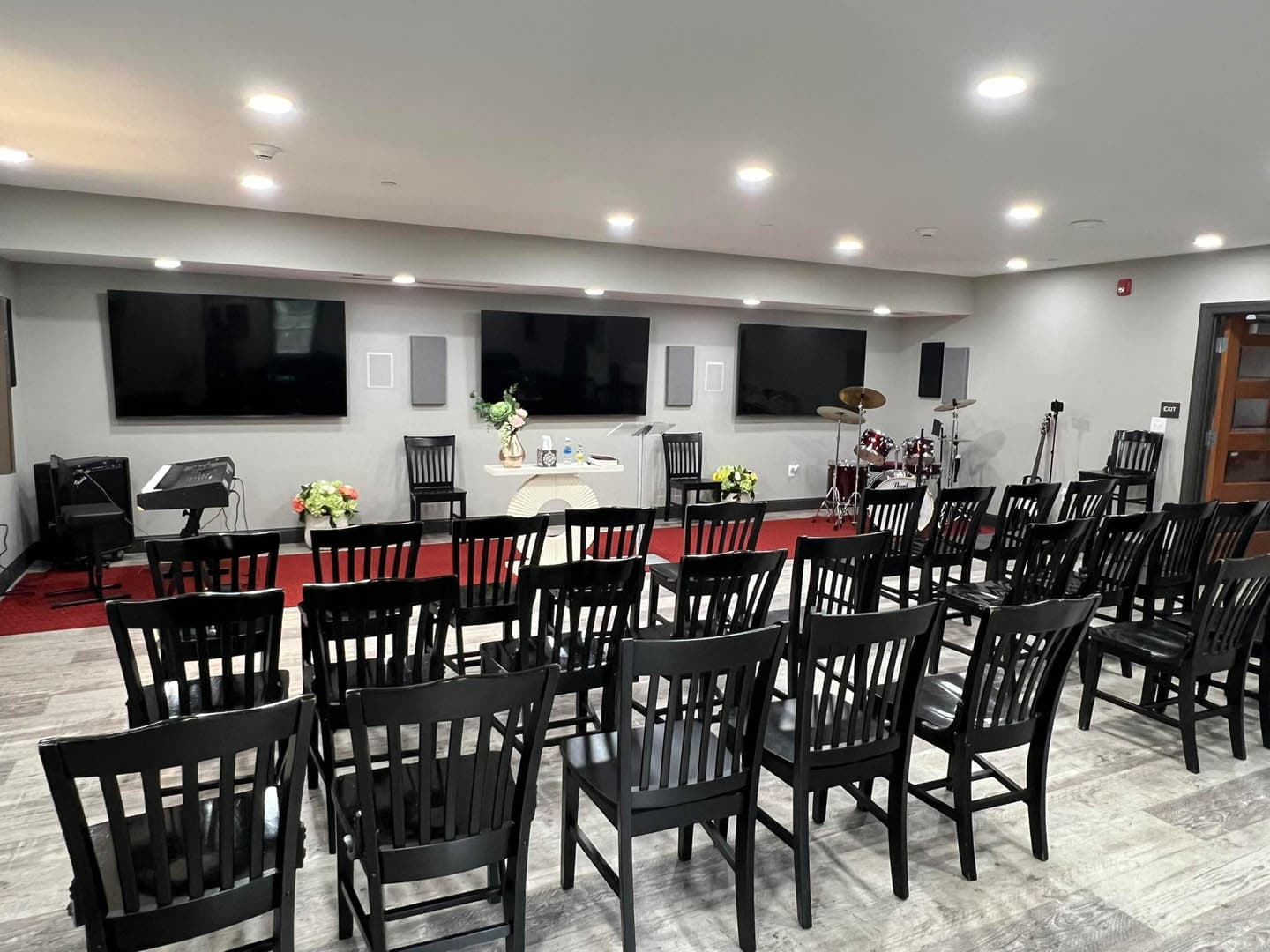 Interior of a completed church worship room with rows of black wooden chairs facing a small altar, drum kit, and multiple large flat-screen televisions mounted on light gray walls