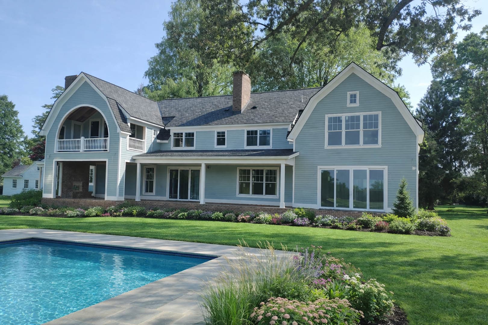 Exterior of a completed two-story light blue shingle-style residence with an arched gable, rear covered porch, swimming pool in the foreground, and mature trees
