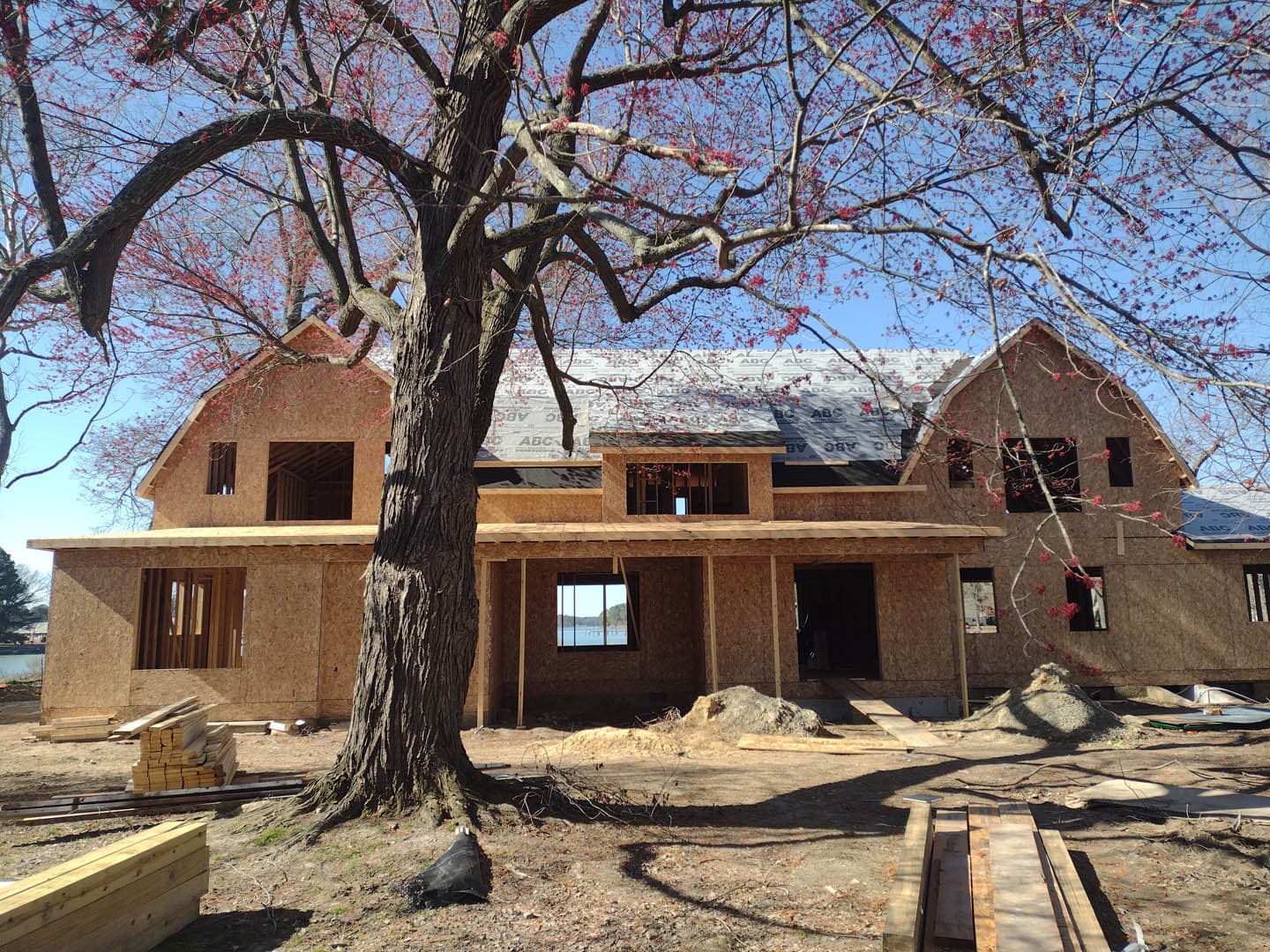 Construction site of a two-story residential home in early wood framing stage with OSB sheathing and partial roofing, set beneath a flowering tree
