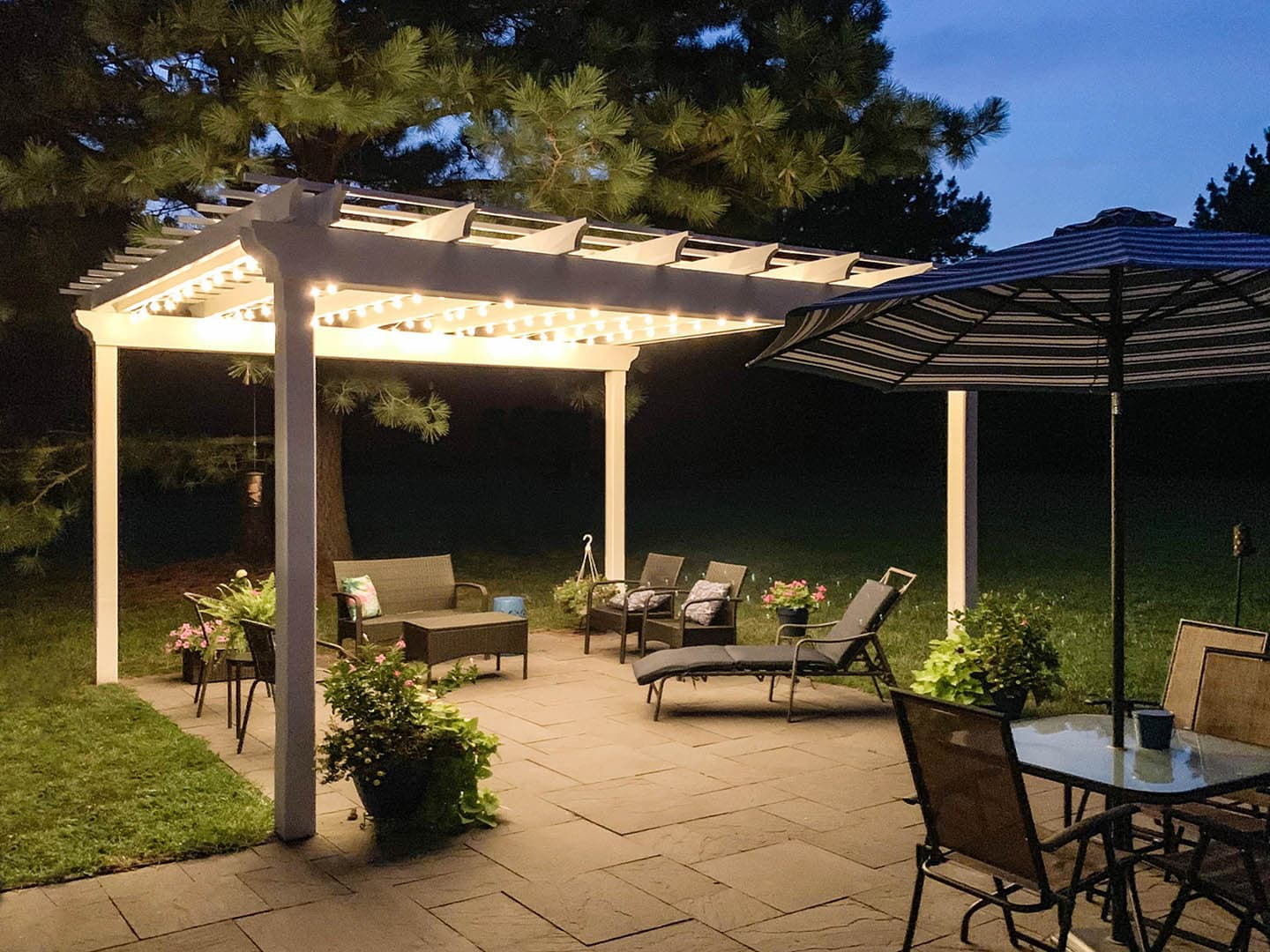 Evening photograph of a white painted pergola over a stone paver patio decorated with warm string lights, surrounded by pines and flowering potted plants