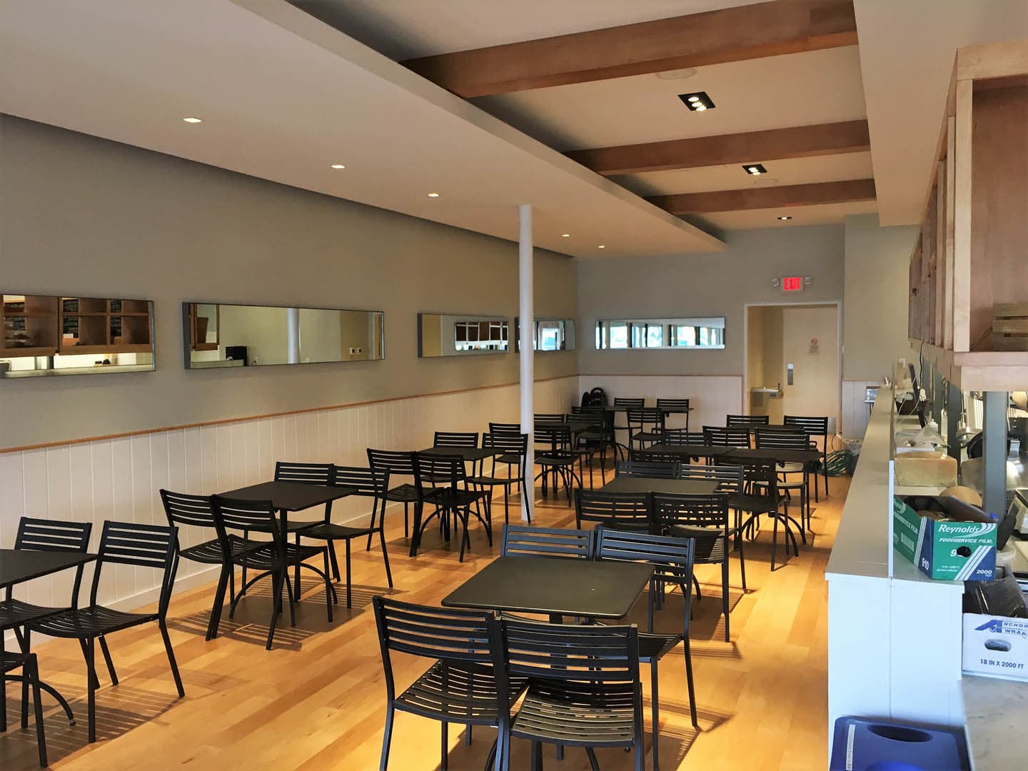 Interior of a completed casual restaurant dining room with light hardwood floors, black metal bistro chairs, white wainscoting, and exposed wood ceiling beams