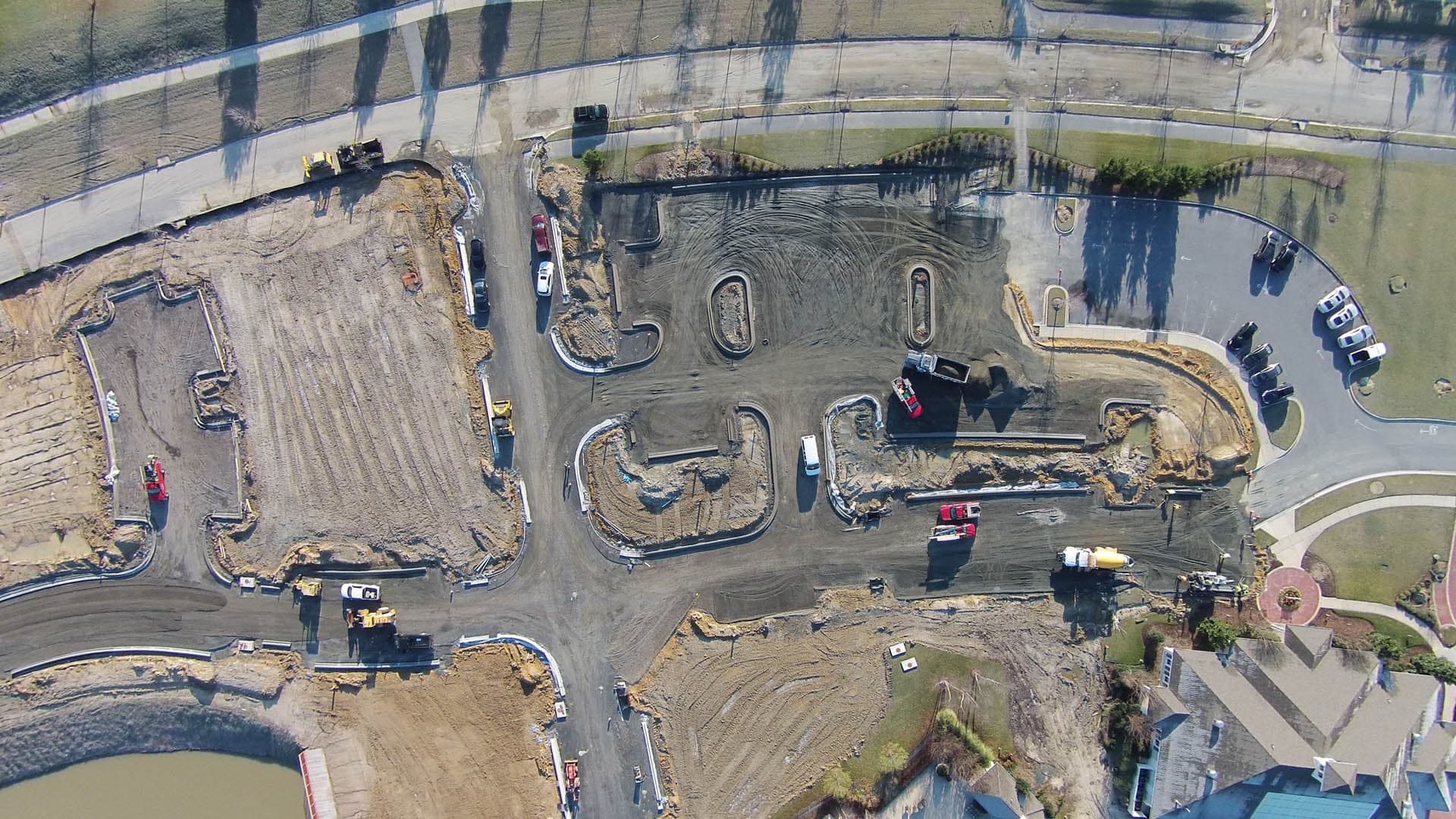 Aerial view of an active road construction site showing utility trench layout patterns in bare soil, construction equipment, and an adjacent pond