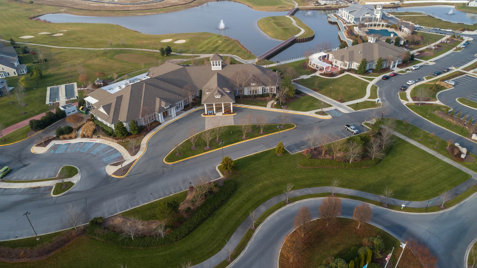 Aerial view of a waterfront community clubhouse with a porte-cochere entrance, curving drive, marina, decorative fountain pond, and golf course