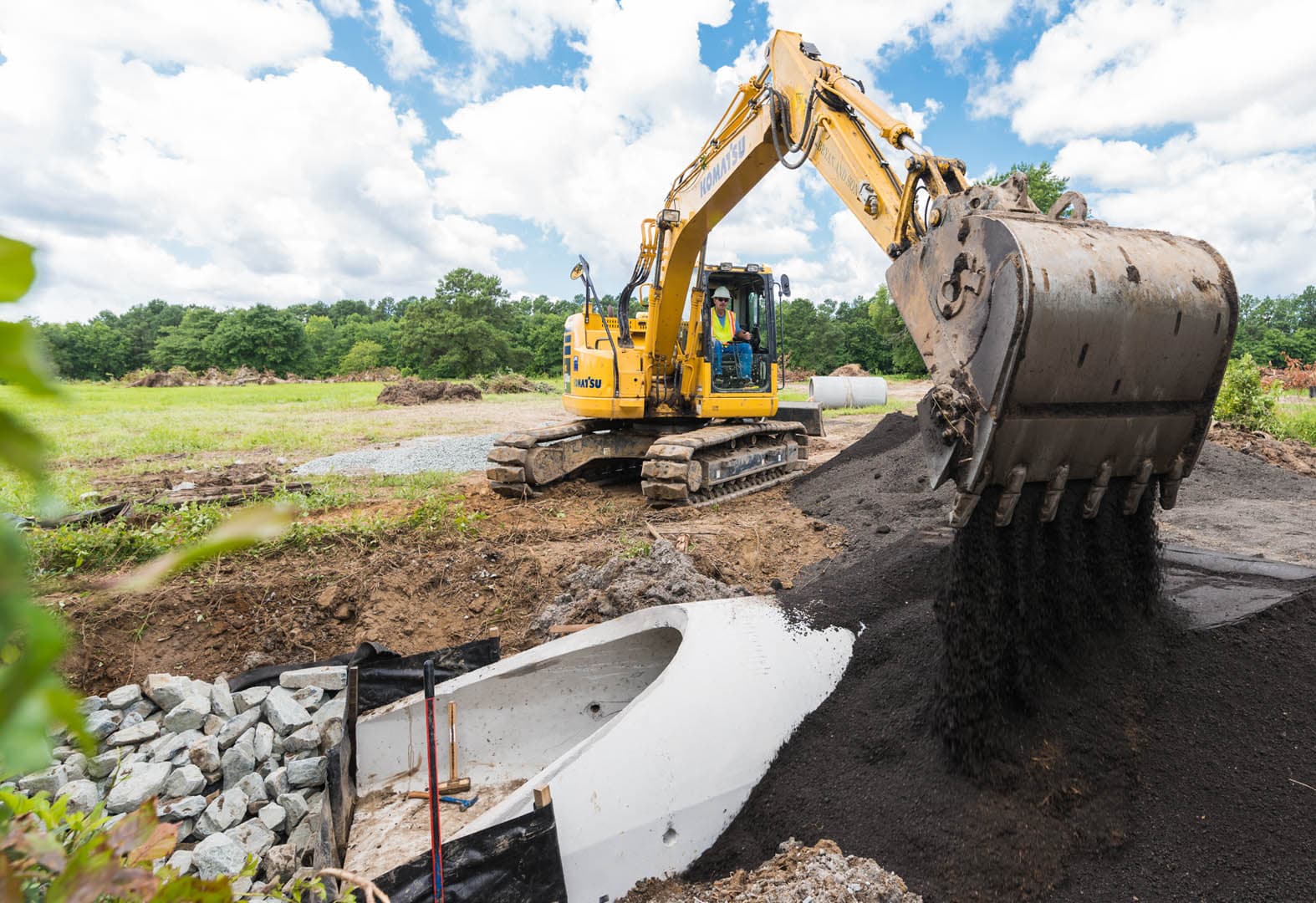 Komatsu excavator dumping soil to backfill a white precast stormwater inlet structure surrounded by rip rap stone