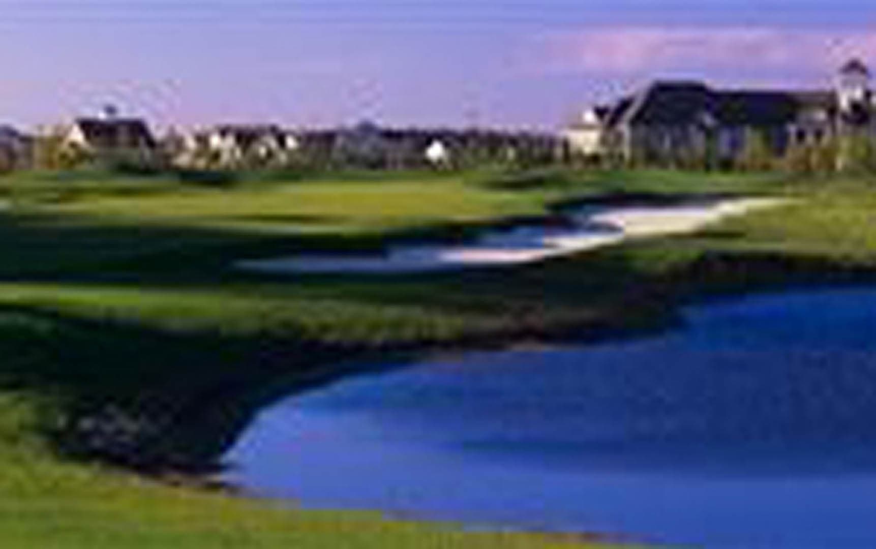 Golf course hole with a large water hazard pond, sand bunker, and manicured fairway, with residential homes visible on the horizon