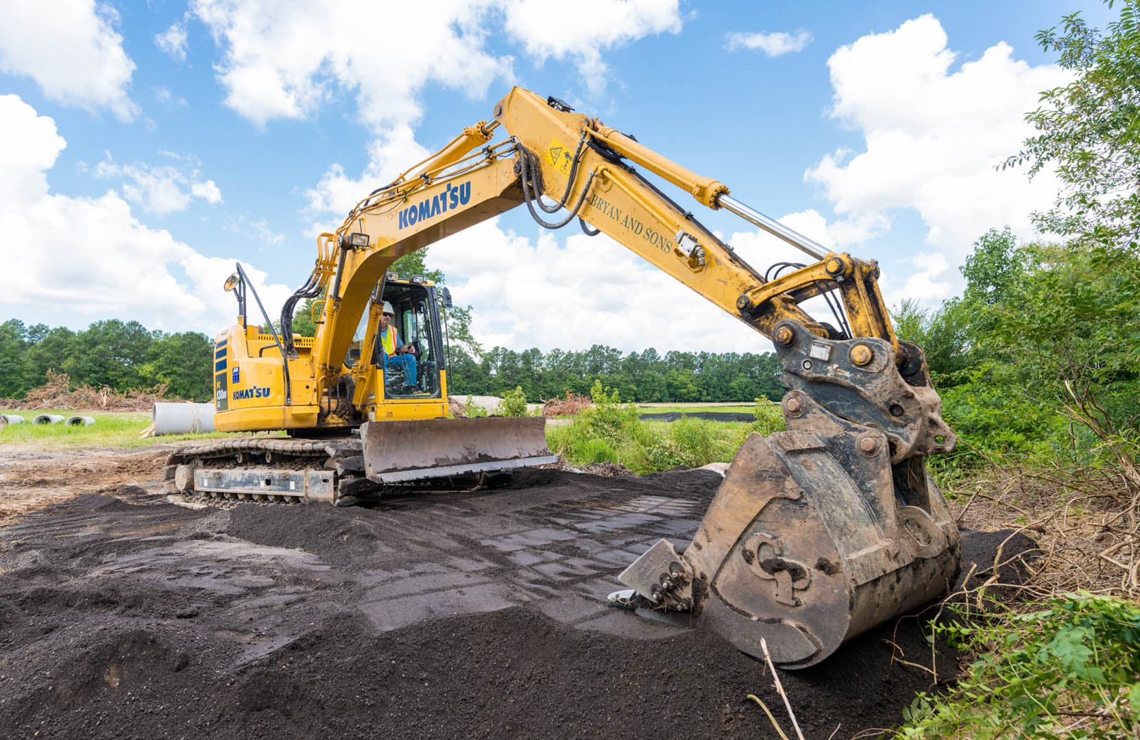 Komatsu excavator grading dark soil on an open construction site with large drainage pipe sections visible in the background