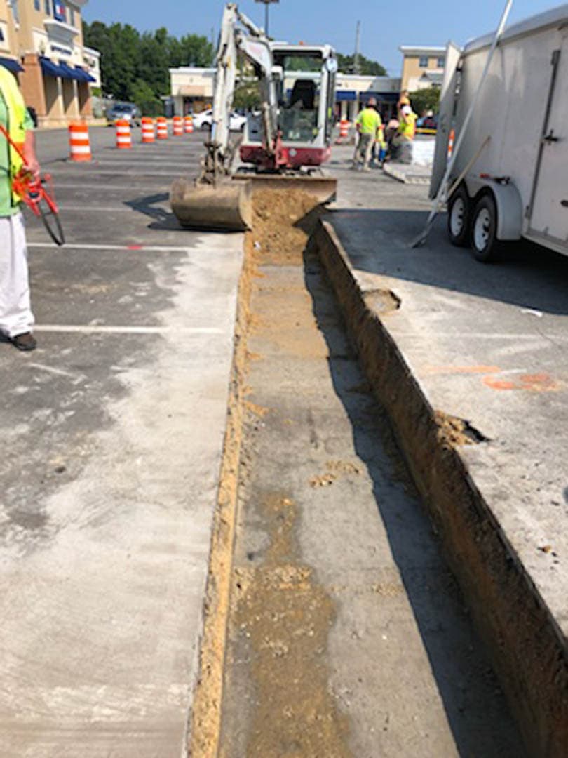 Mini excavator cutting a narrow utility trench through an asphalt parking lot with traffic control barrels and workers in high-visibility vests