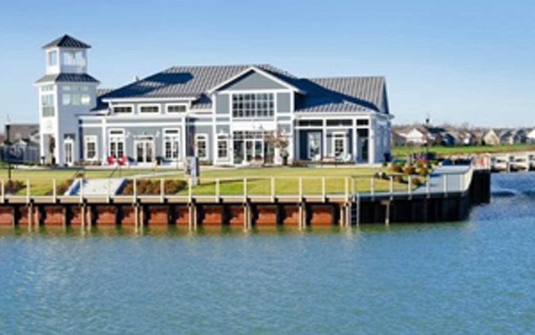 Ground-level view of a large waterfront clubhouse with gray siding and standing seam metal roof, seen from across the water with a timber dock and boardwalk