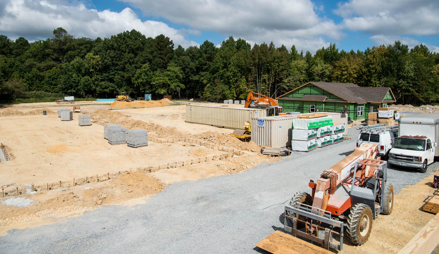 Wide-angle view of a commercial building construction site showing concrete block foundation footings, a telescopic forklift, and stacked building materials