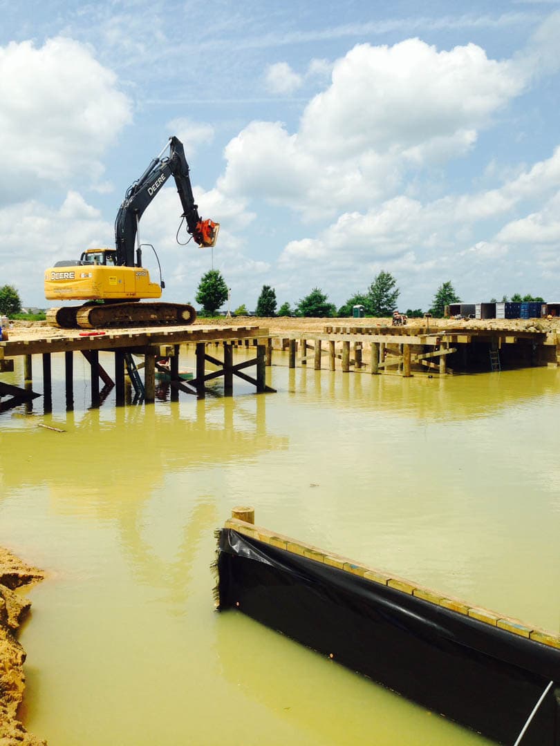 John Deere excavator with vibratory hammer attachment driving timber piles into a flooded construction basin from an elevated work platform