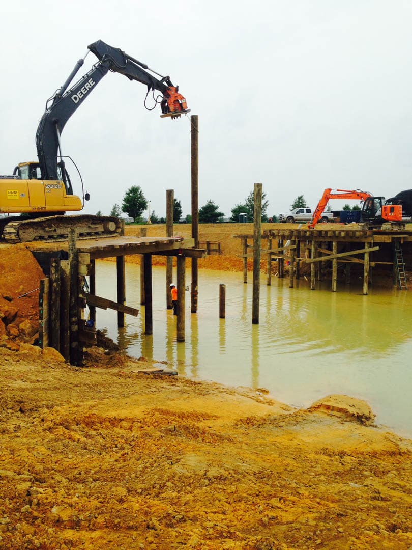 Two excavators working in tandem driving timber piles into a flooded site, with a worker in high-visibility gear standing between the pile rows