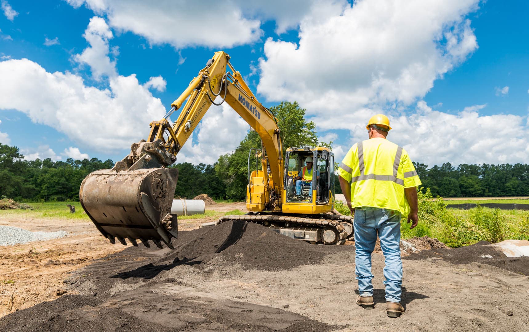 Construction inspector in yellow high-visibility vest and hard hat standing near a Komatsu excavator moving fill material on an active earthwork site