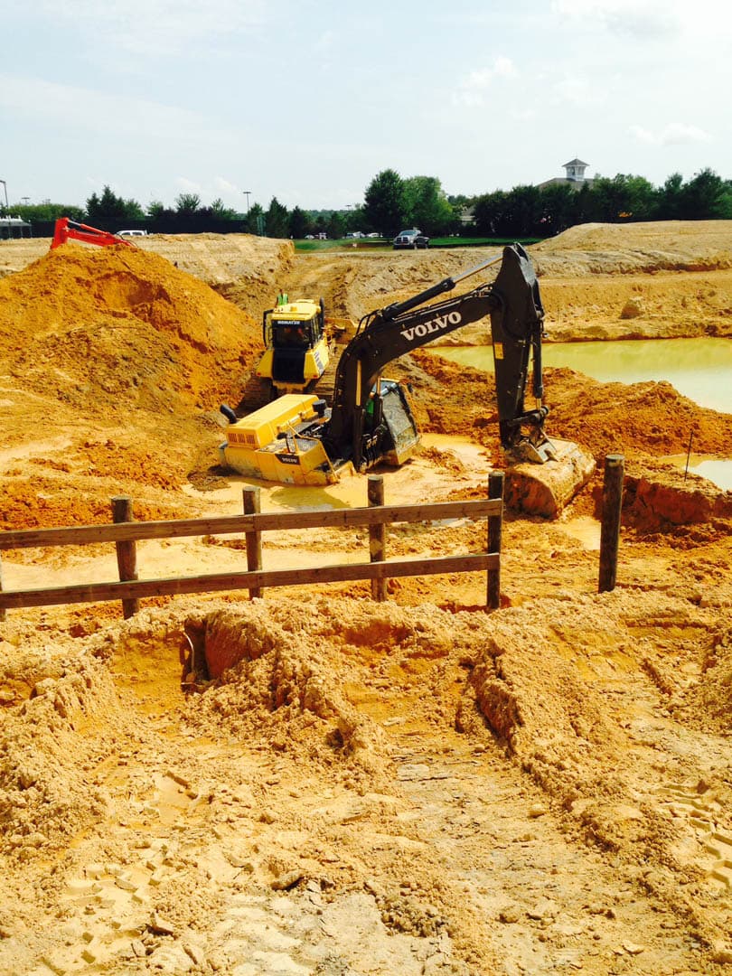 Volvo excavator digging near a retention pond during earthwork excavation, with sandy soil mounds and wooden stake markers