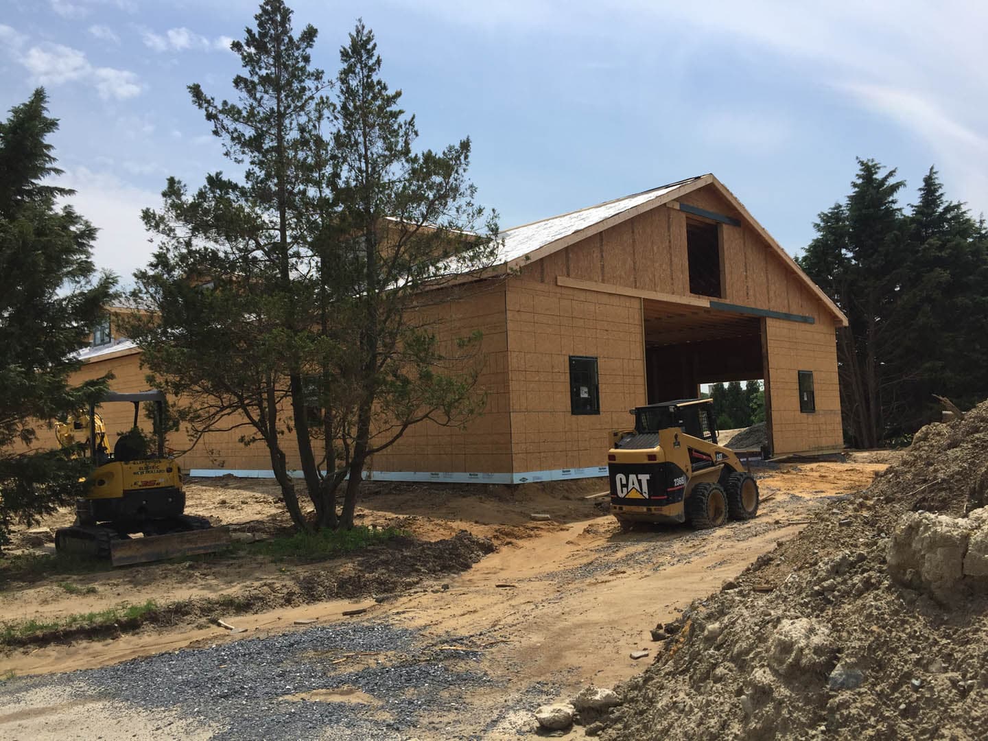 Newly framed wood structure with OSB sheathing and metal roofing partially installed, with a CAT skid steer loader and mini excavator grading the site