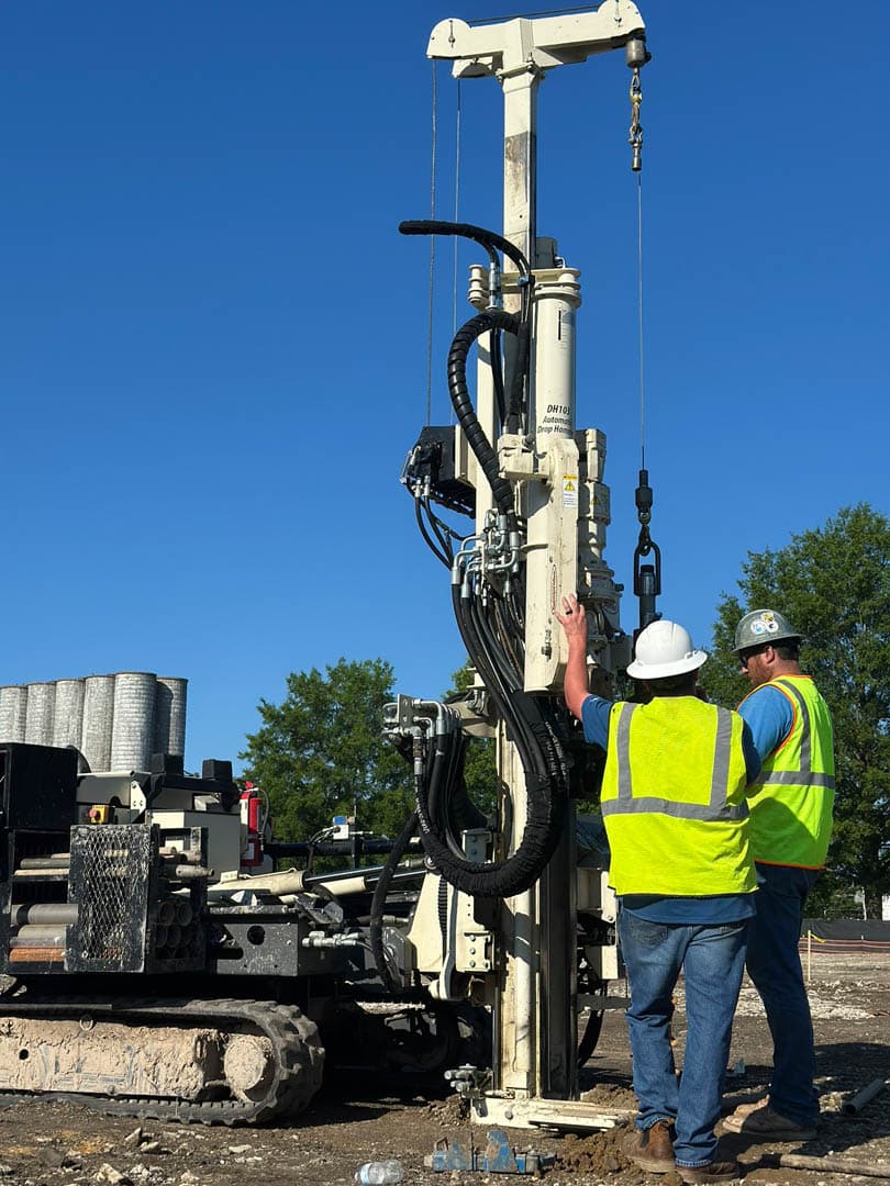 Environmental worker in yellow safety vest and hard hat operating a tall track-mounted direct-push geoprobe drill rig against a clear blue sky at an industrial brownfield site