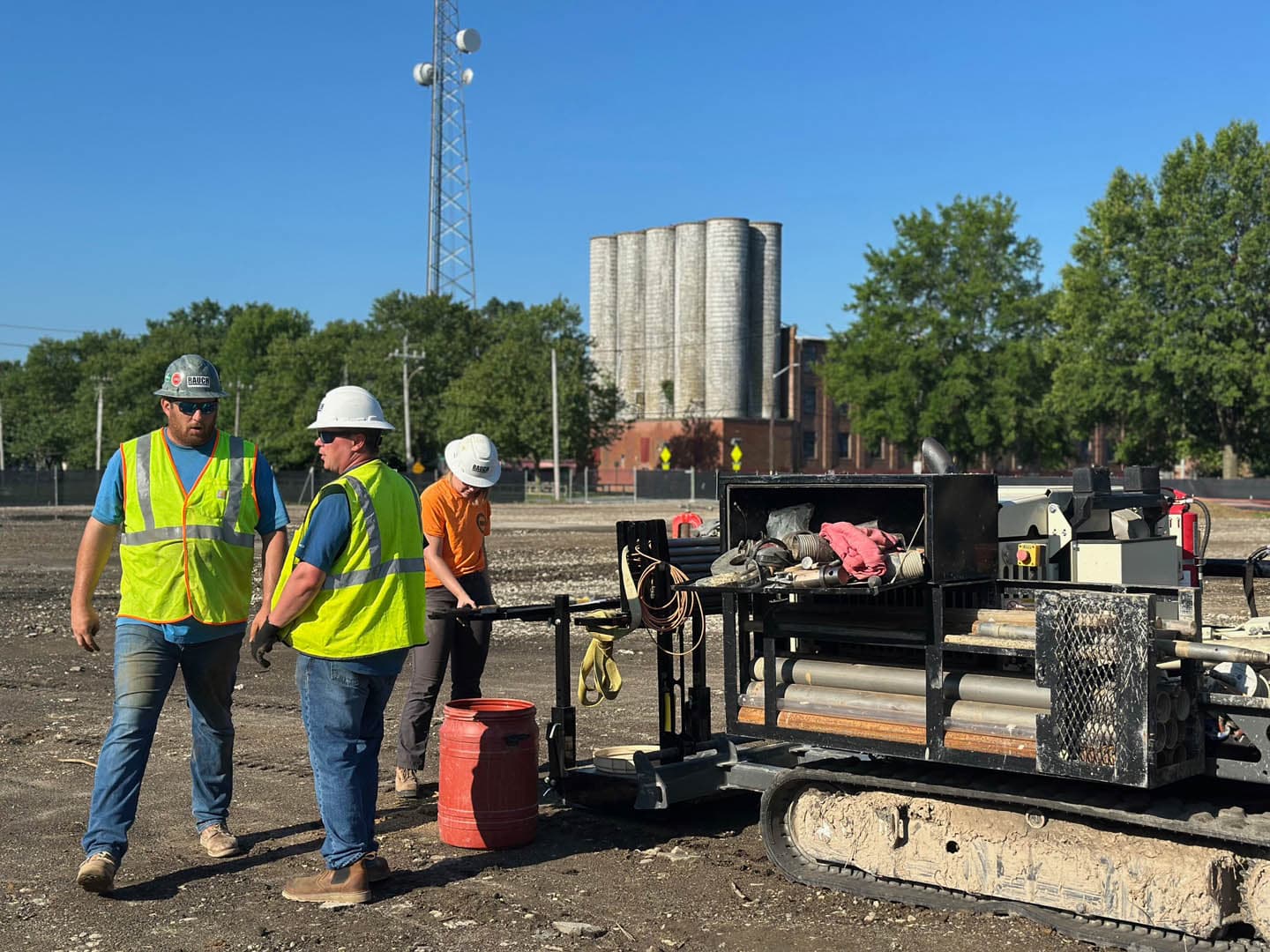 Three-person environmental drilling crew in safety vests and hard hats working with a track-mounted direct-push drill rig and support equipment at an urban brownfield site with grain silos in the background