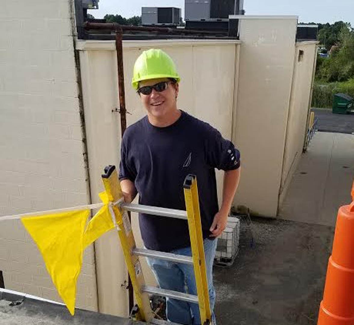 Male environmental inspector in a yellow hard hat and sunglasses standing on a ladder against the exterior wall of a commercial building during a facility inspection