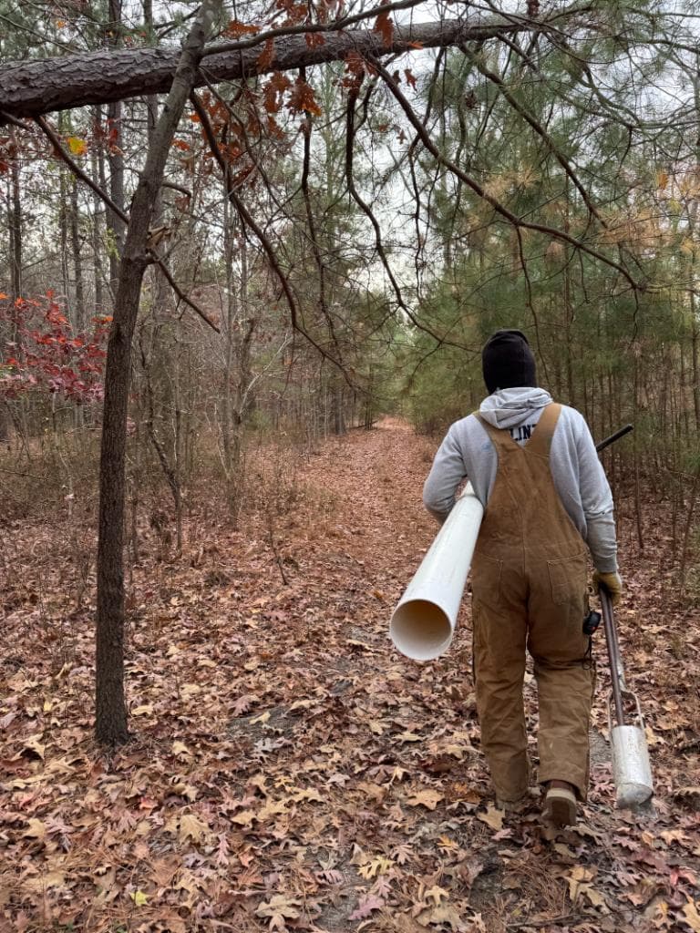 Environmental field technician in overalls and winter hat walking a forest trail carrying white PVC well casing pipe for groundwater monitoring well installation