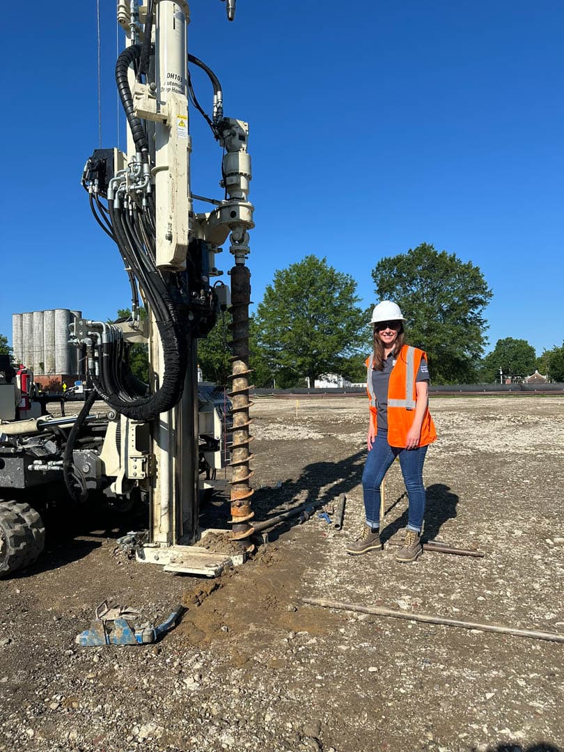 Female environmental scientist in orange safety vest and white hard hat standing beside a direct-push drill rig with exposed auger bit during soil investigation at a brownfield site