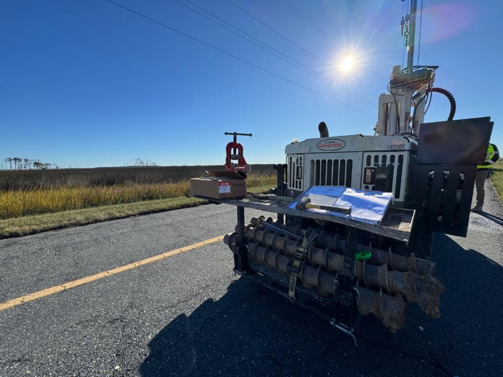 Geoprobe 7822DT direct-push drilling rig staged on a rural road adjacent to coastal marshland under a bright blue sky