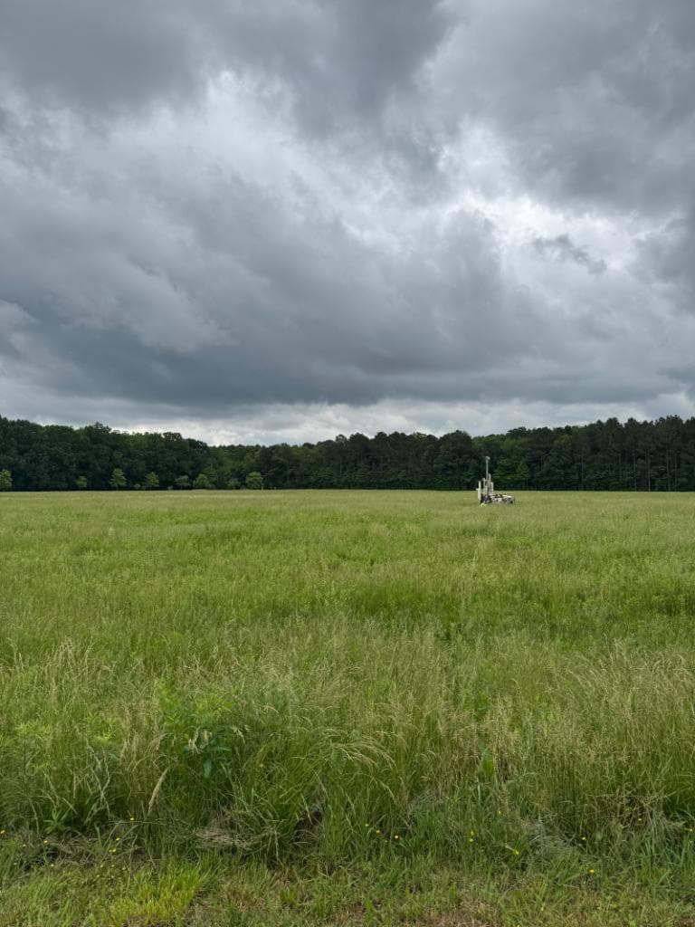 Geotechnical drilling rig standing in a wide open grassy field under dramatic storm clouds with a forested treeline in the background