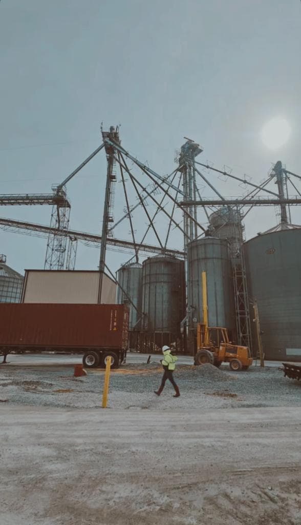 Environmental professional in white hard hat and safety vest conducting a site assessment at an industrial grain storage facility with large silos and conveyors