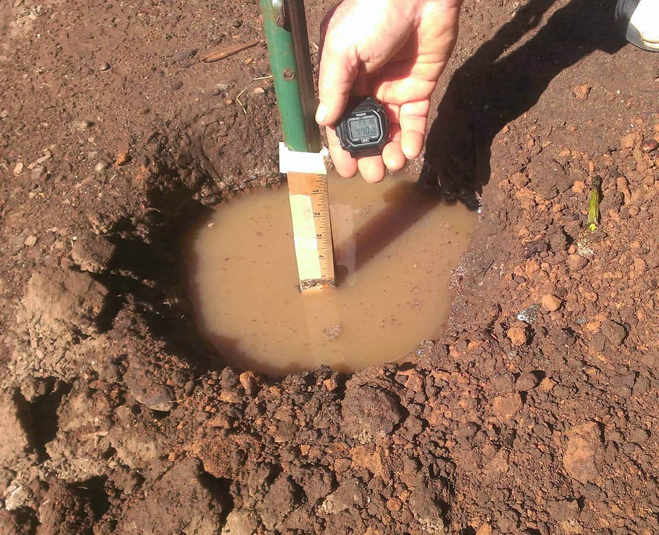 Close-up of a hand holding a stopwatch over a ruler measuring water level in a soil percolation test hole during a site evaluation for septic system design