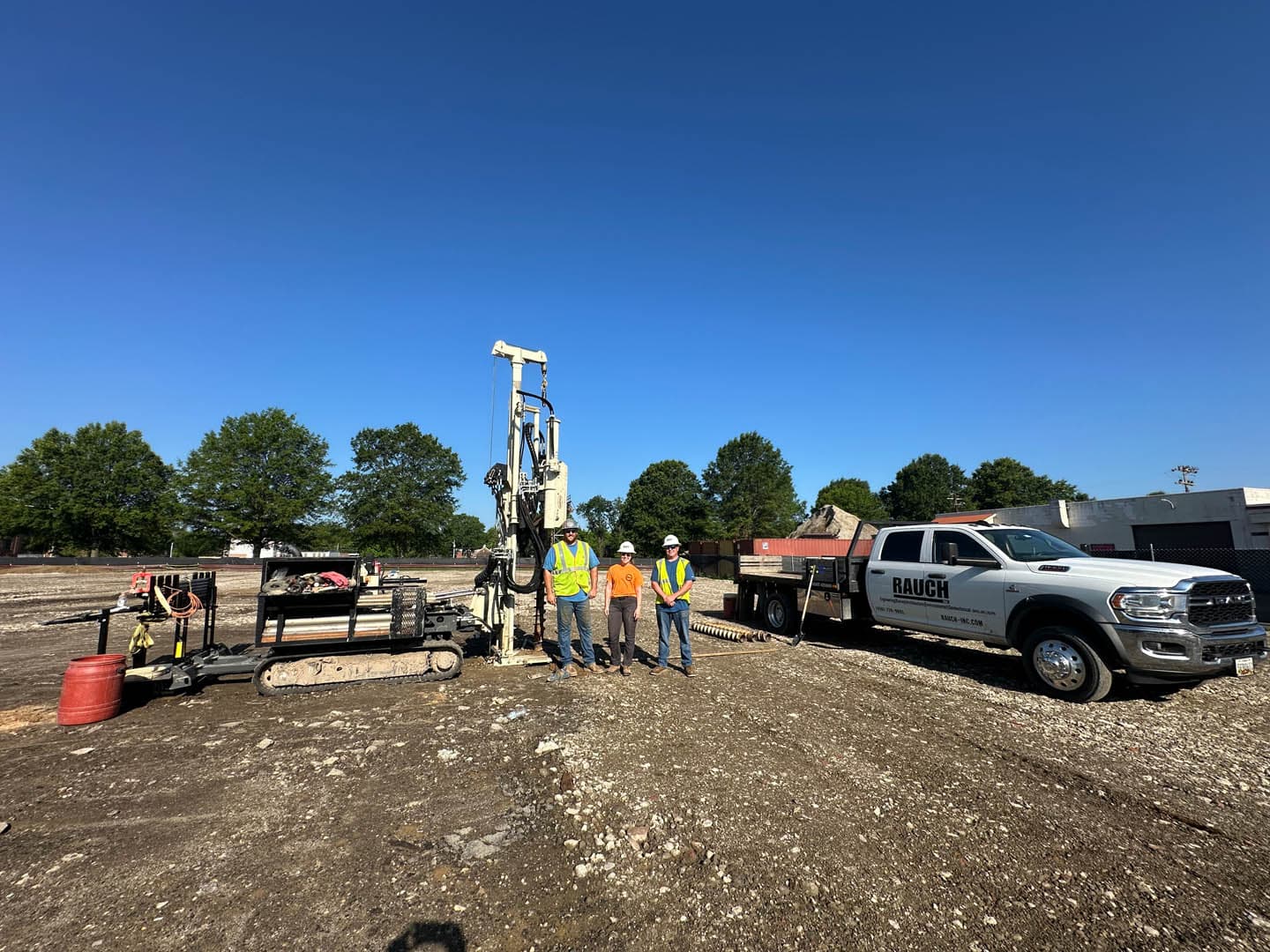Three RAUCH environmental team members in safety vests and hard hats posing in front of a direct-push drill rig and a white RAUCH-branded pickup truck at a brownfield investigation site
