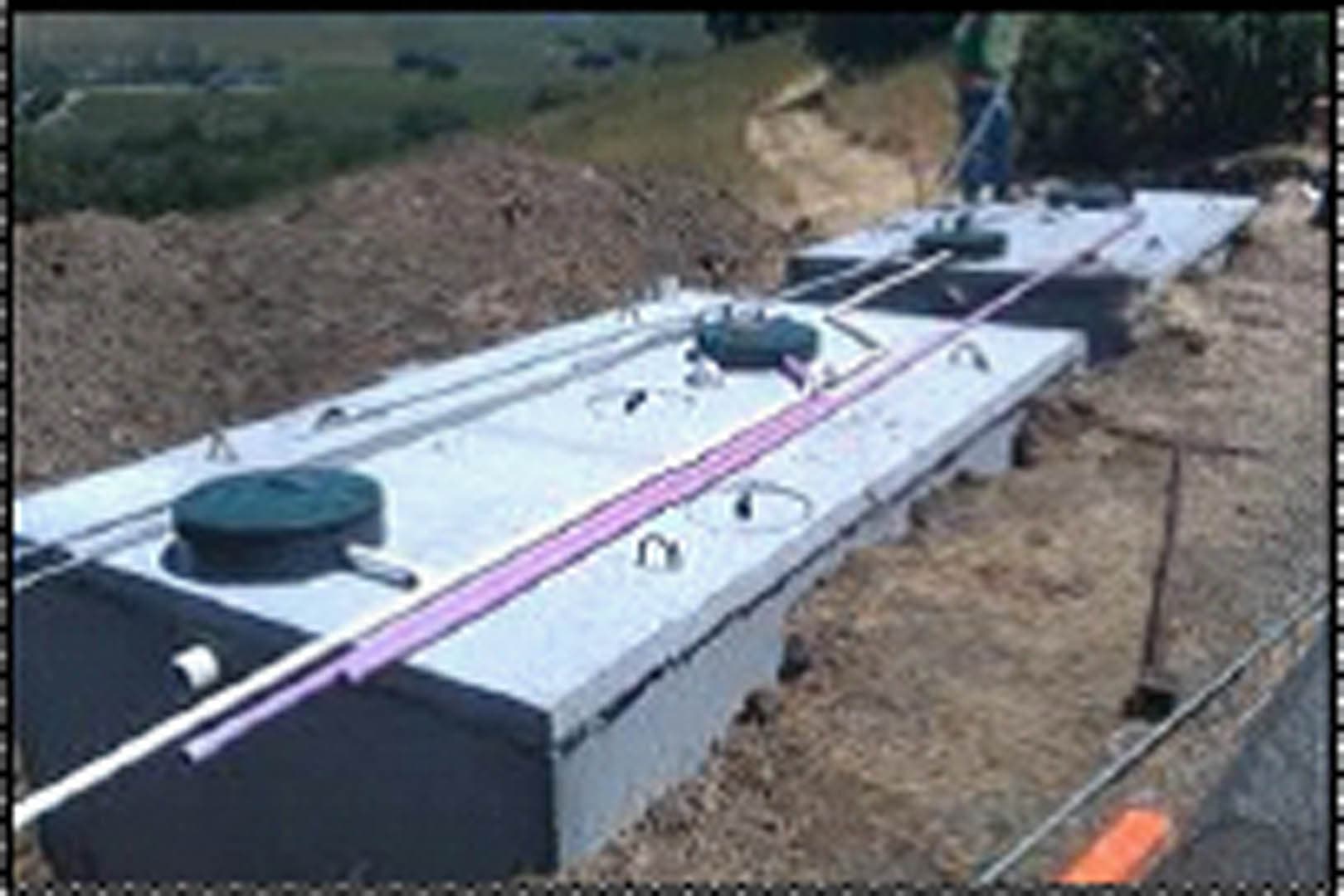 Aerial view of a large concrete septic tank system installed in an excavated hillside with access lids, piping, and purple distribution lines visible on the tank surface