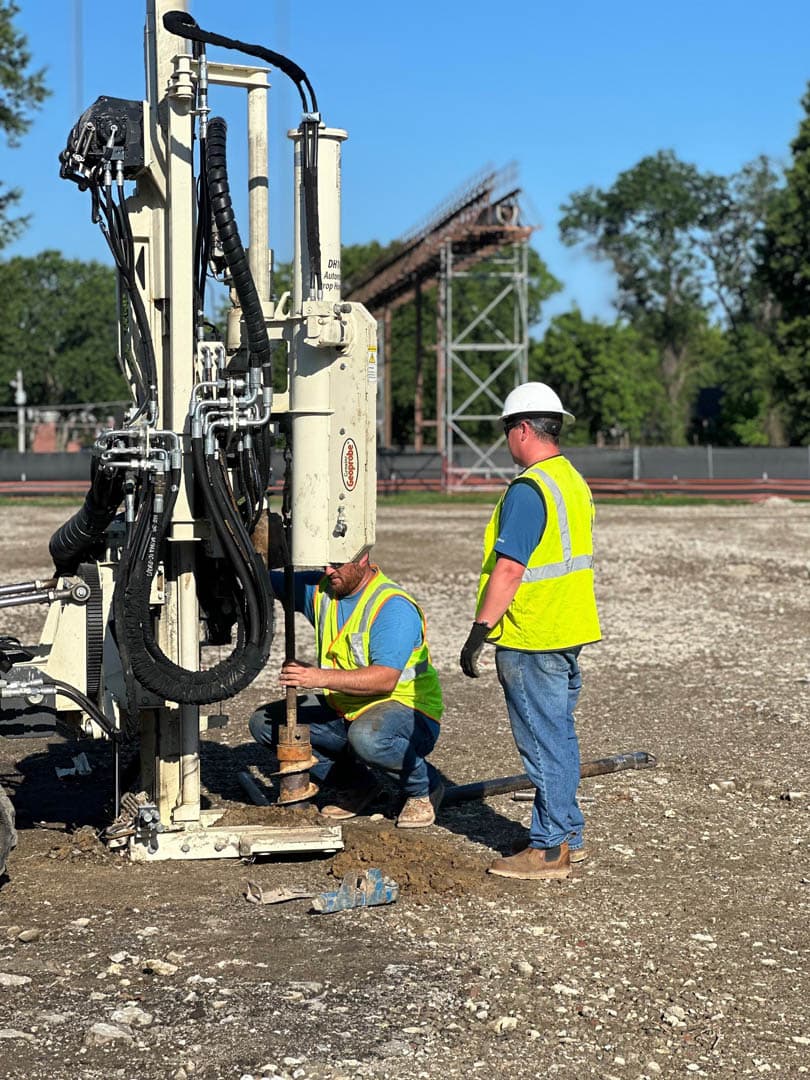 Two environmental workers in high-visibility vests and hard hats crouching beside a soil boring drill rig operating at a brownfield site