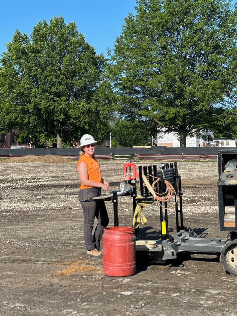 Environmental field technician in orange safety vest and white hard hat standing at a soil boring drill rig on a cleared gravel site