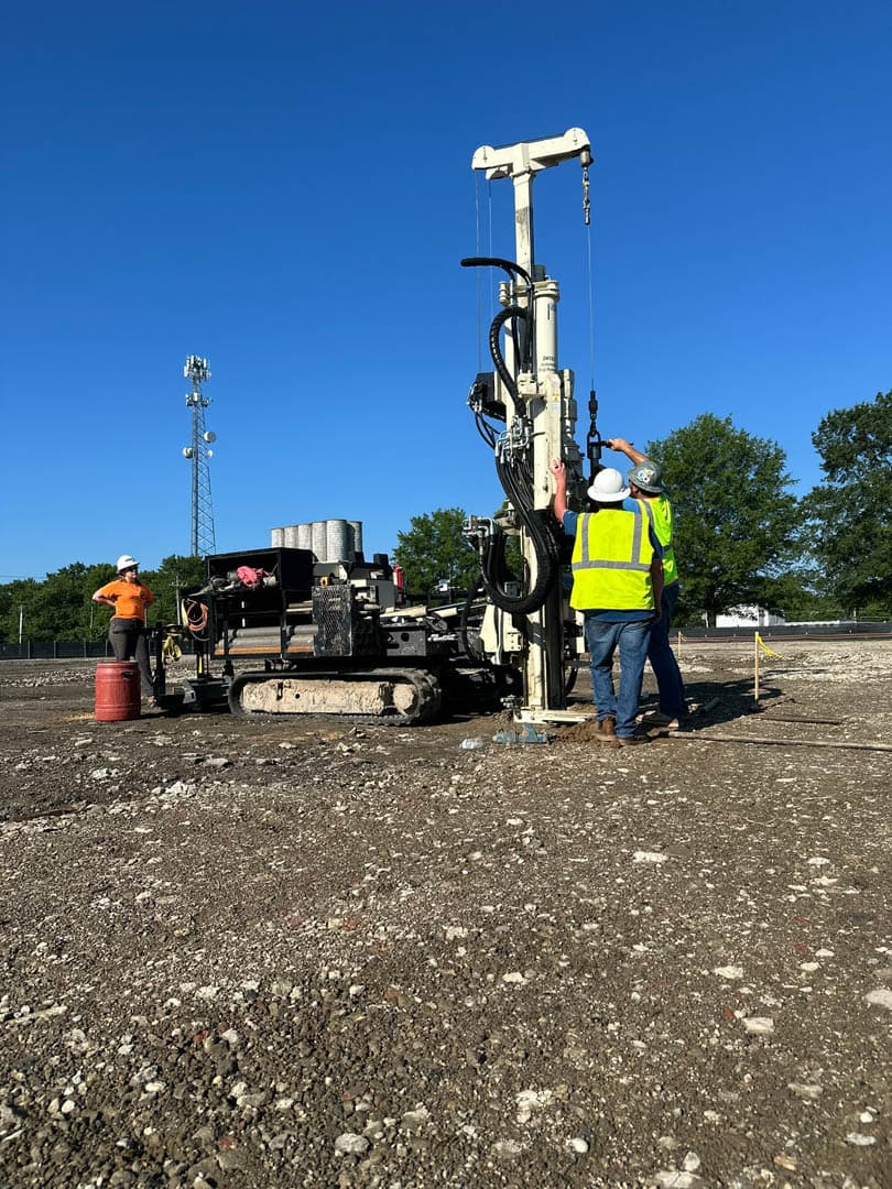 Wide shot of two workers and a track-mounted drill rig conducting soil sampling at a gravel brownfield site with a communications tower and industrial buildings in the background