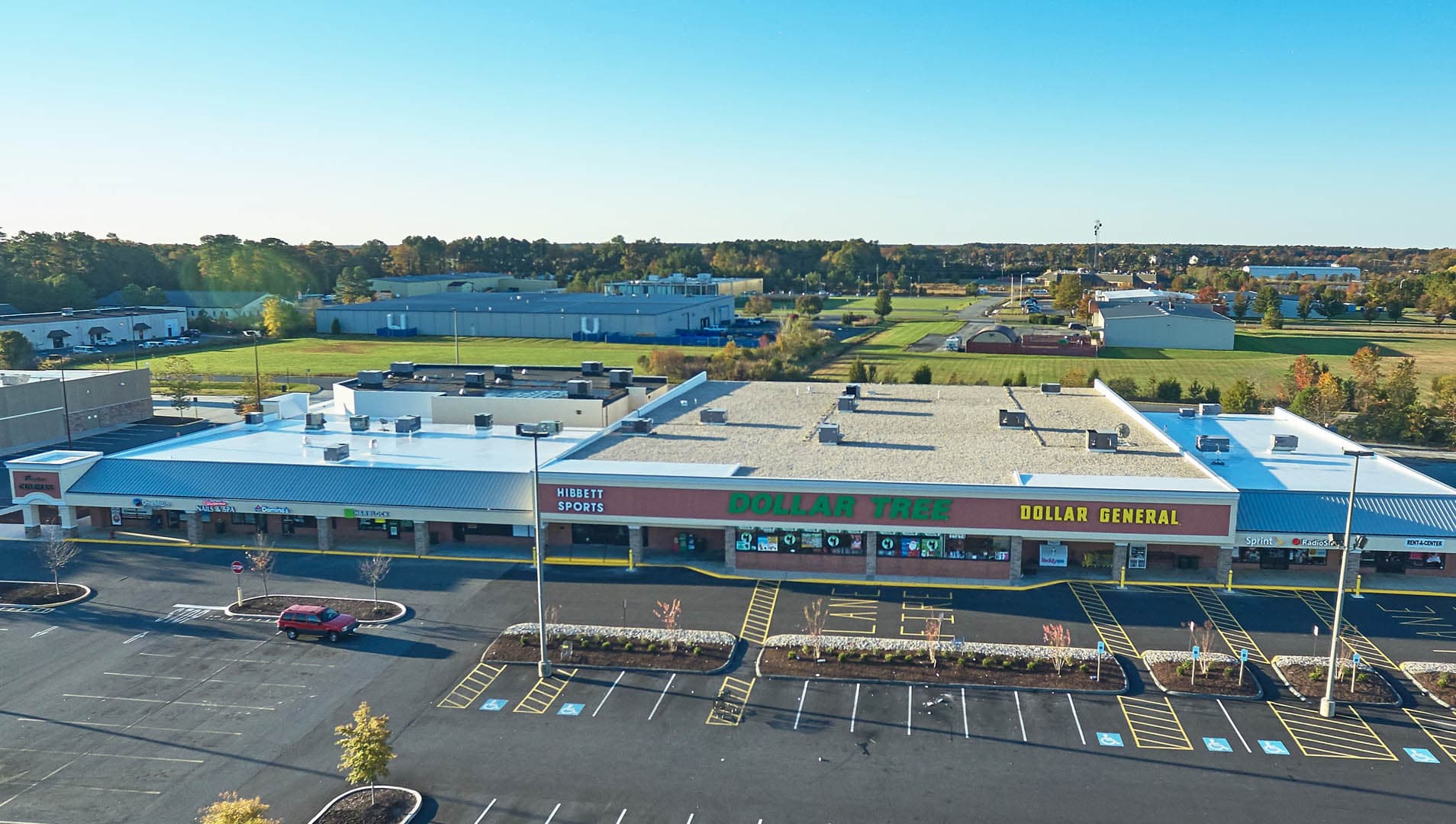 Aerial drone photograph of a commercial strip mall with storefronts, a large paved parking lot, and surrounding industrial buildings