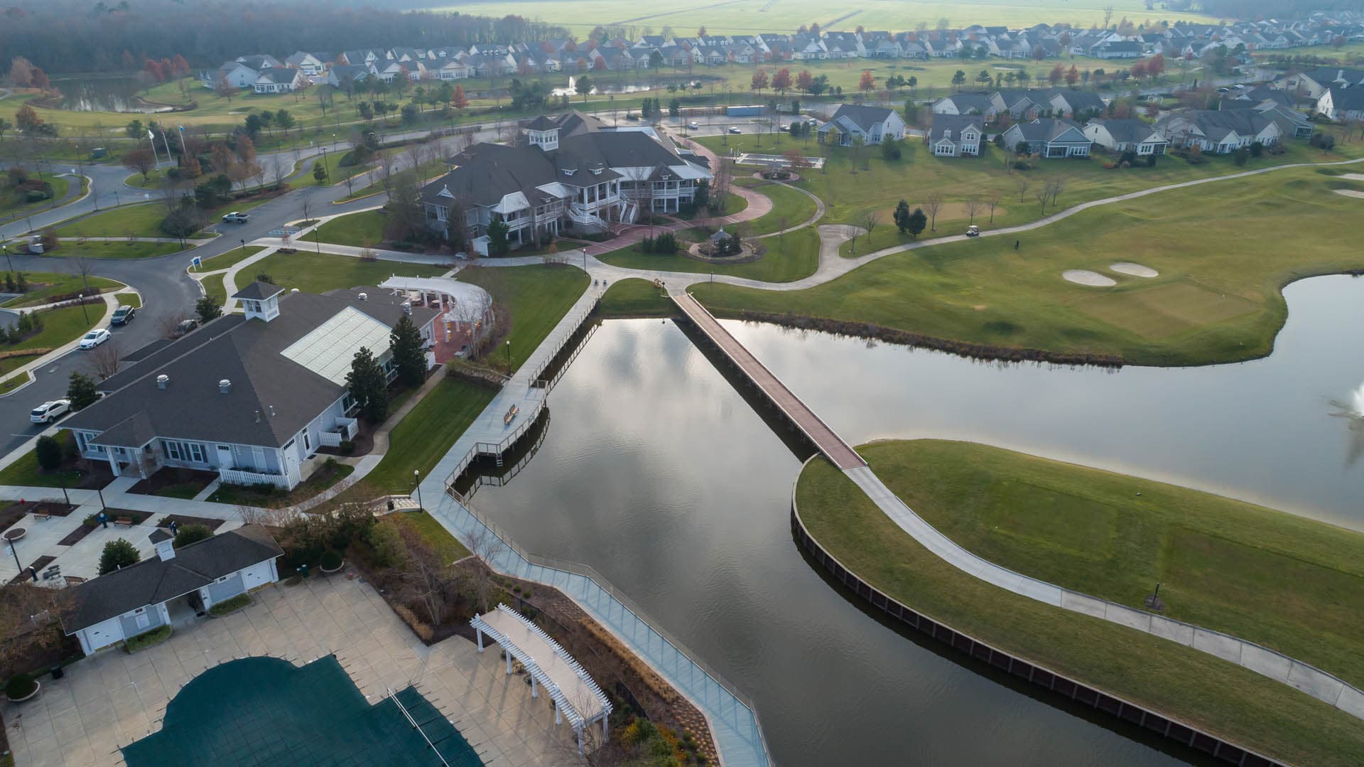 Aerial drone photograph of a residential golf course community showing a large clubhouse, swimming pool, pedestrian bridge over a stormwater pond, and surrounding homes