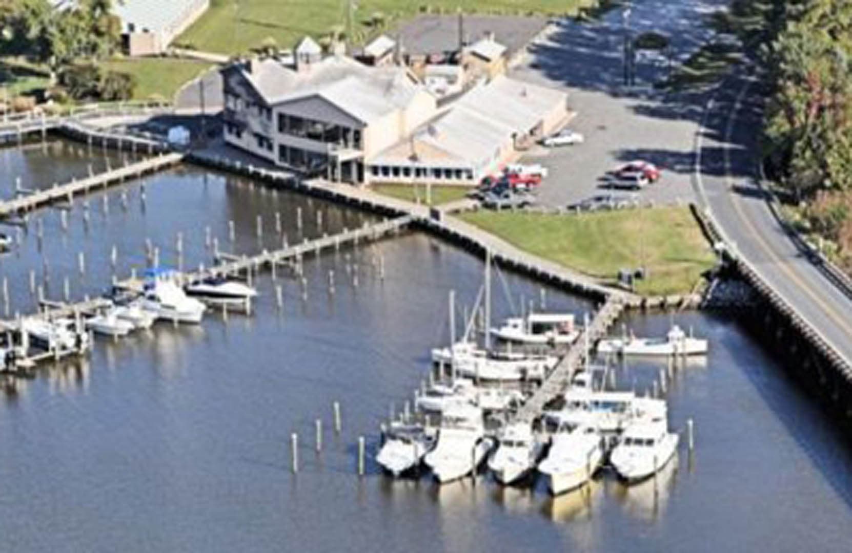 Aerial photograph of a small marina on a tidal waterway showing multiple boat slips with docked sailboats and powerboats and a waterfront building complex