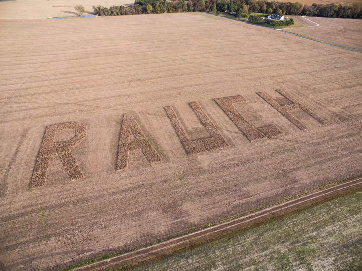 Aerial drone photograph of a harvested farm field with the word RAUCH carved in large block letters into the crop stubble