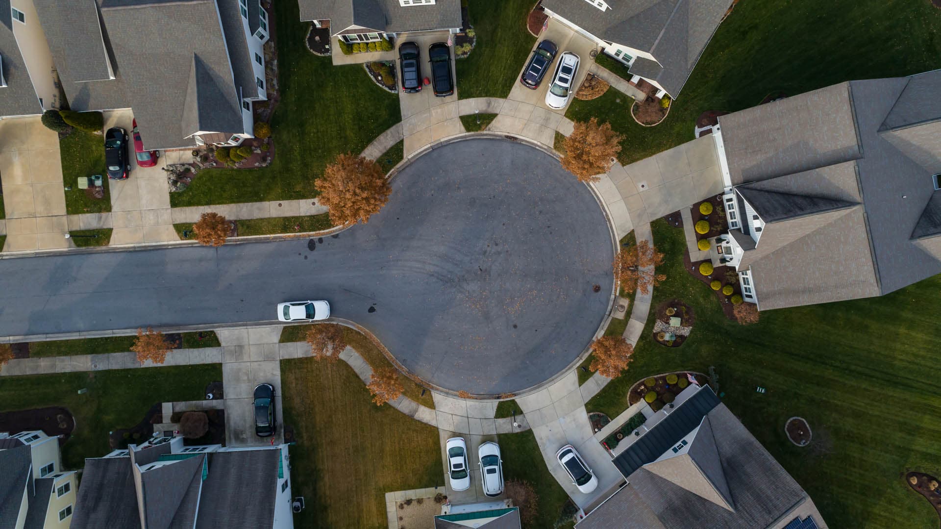Straight-down aerial drone photograph of a residential cul-de-sac with driveways, parked vehicles, sidewalks, and landscaped yards in an autumn neighborhood