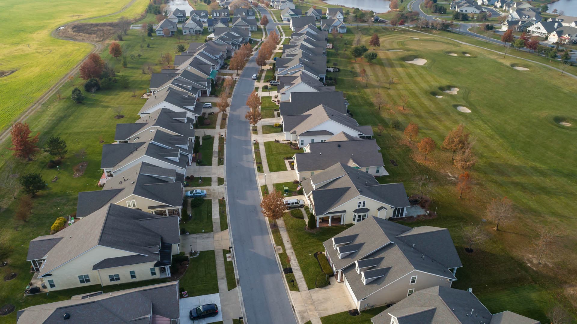 Aerial drone photograph of a residential subdivision street lined with single-family homes in autumn, bordered by a golf course with sand traps and waterfront