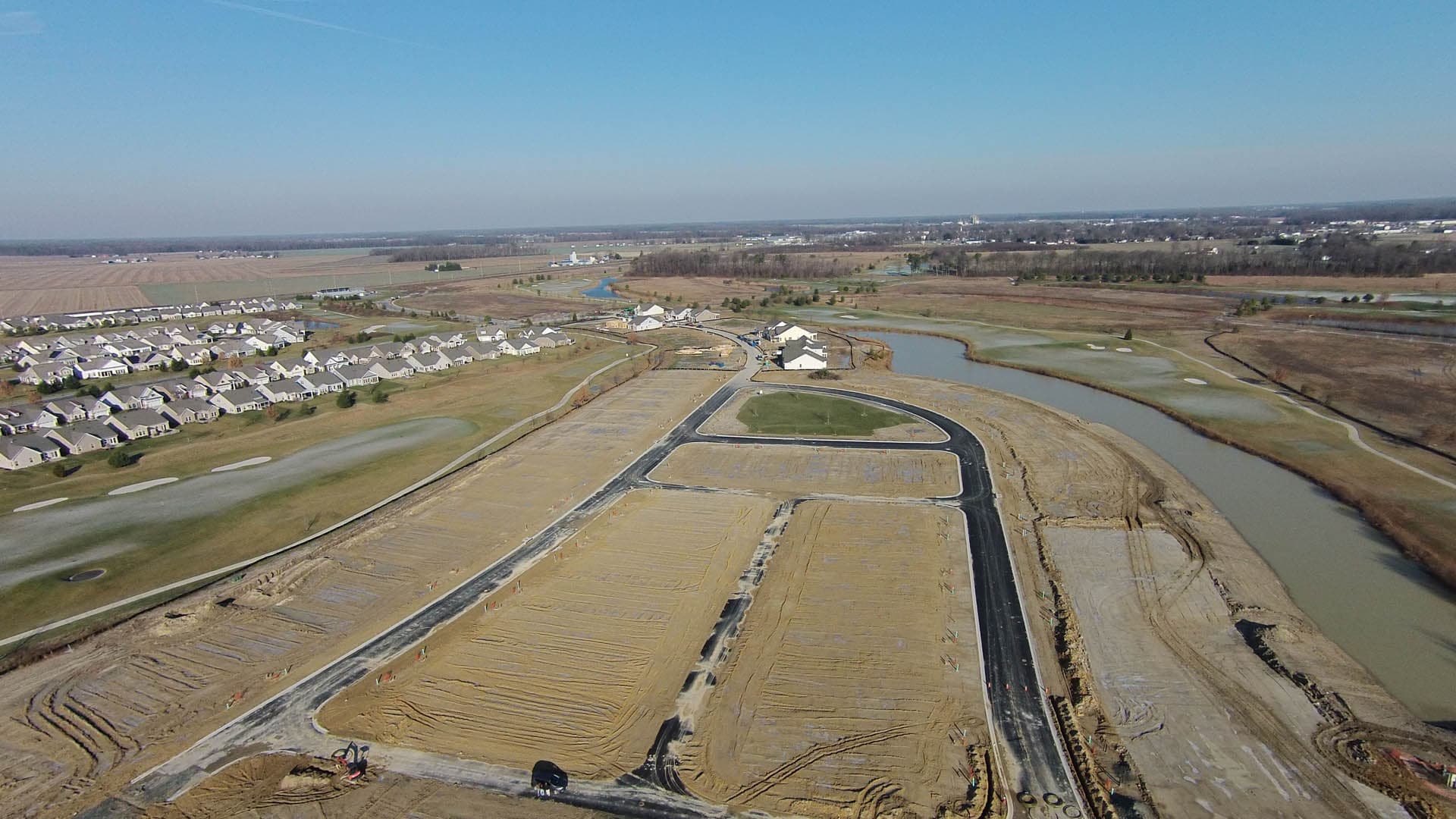 Wide aerial drone photograph of a large residential subdivision under construction showing graded lots, new road infrastructure, and a curving waterway