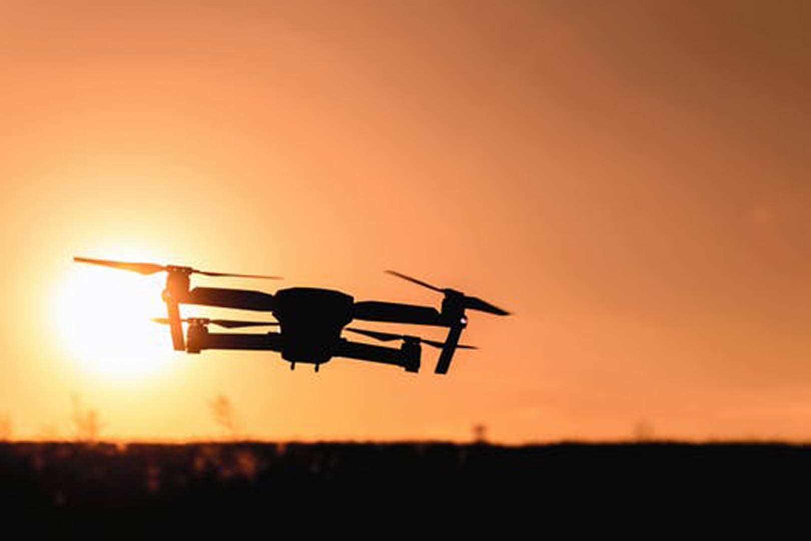 Silhouette of a compact folding drone in flight against a warm golden-orange sunset sky with the sun visible near the horizon