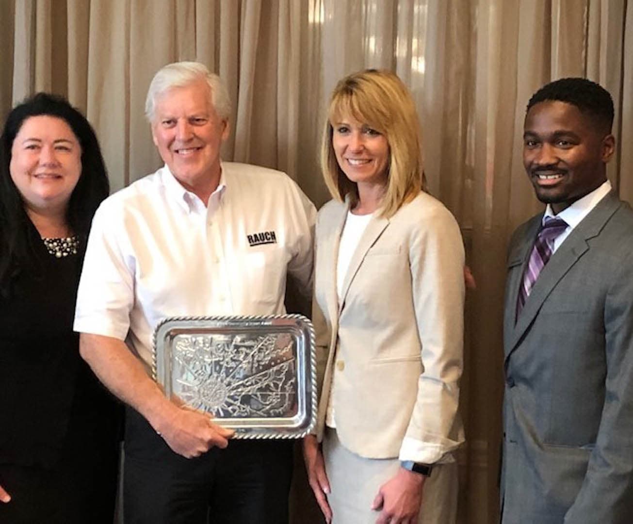 Four people standing together at a formal reception, with a RAUCH Inc. representative holding a silver engraved award platter