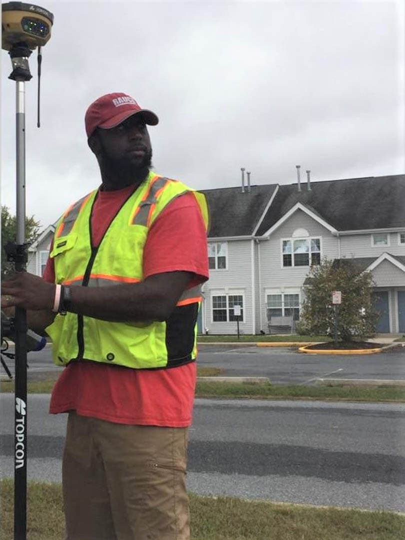 RAUCH survey technician in high-visibility safety vest and red RAUCH cap holding a Topcon GPS rover rod in a parking lot with a residential building in the background