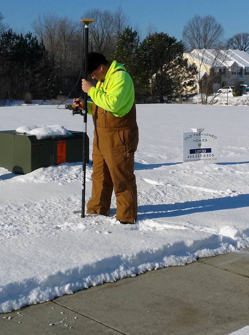 Survey technician in high-visibility hoodie operating a Topcon GPS rover rod in a snow-covered residential lot