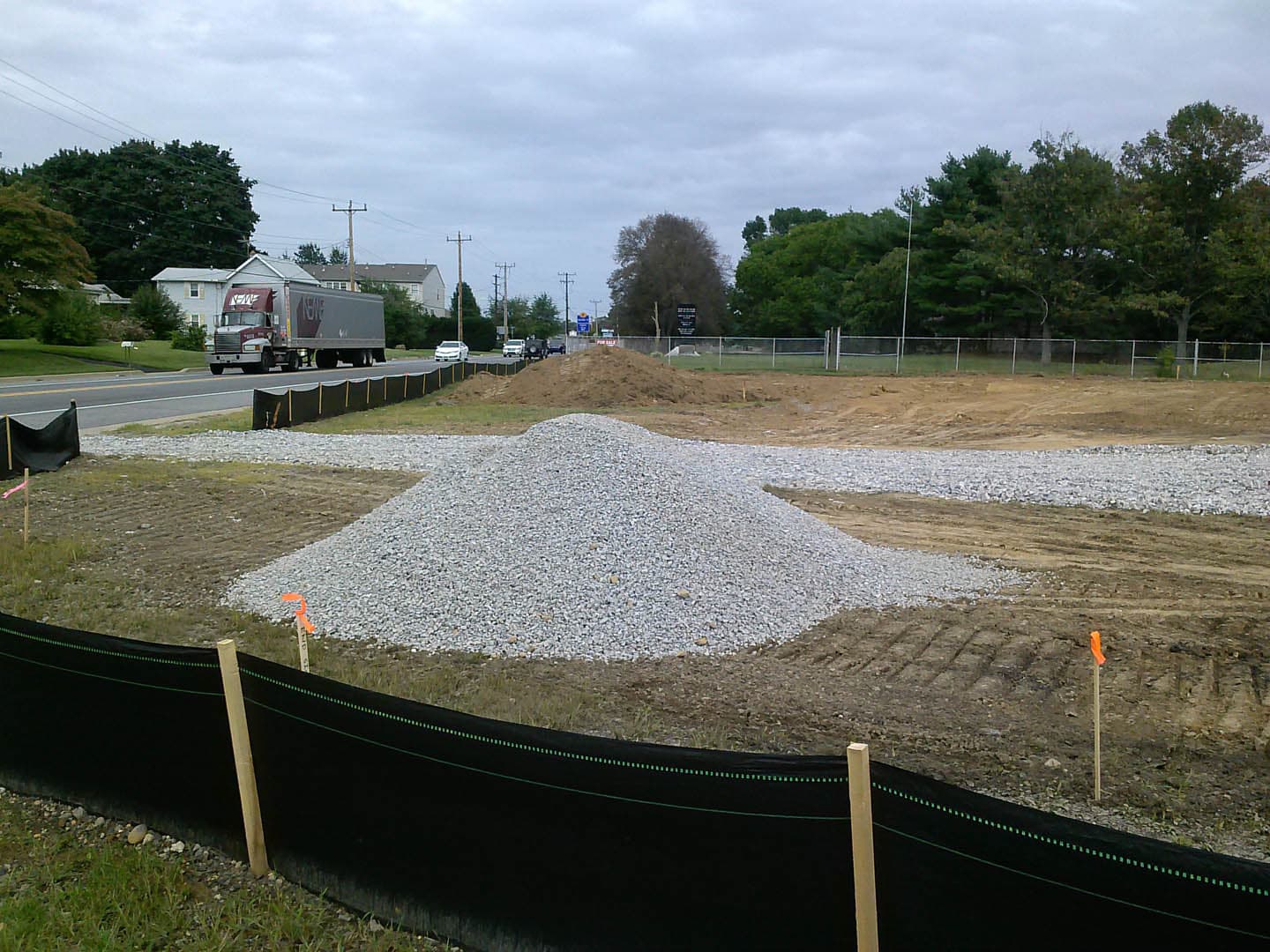 Active construction site with piles of crushed gravel, a silt fence along the roadside perimeter, and orange survey flags marking points